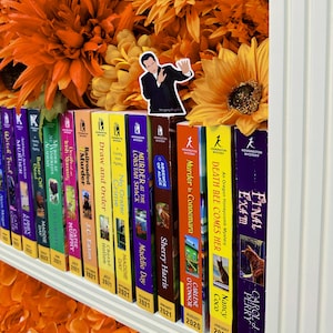May include: A bookshelf filled with colorful paperback mystery novels. The books are arranged by author and publication date. The titles include "Murder at the Lobster Shack", "No Grater Crime", "Draw and Order", "Railroaded", "Death Bee Comes Her", "Final Exam", and "Murder in Connemara".