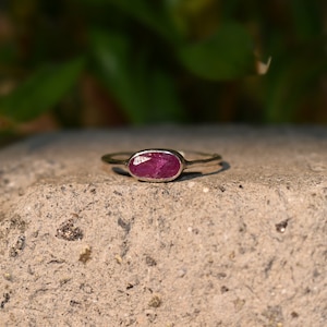 Op de afbeelding: Een zilveren ring met een ovale robijn edelsteen. De ring ligt op een getextureerd, grijs oppervlak, met een vage groene achtergrond. De robijn heeft een diepe, rijke rode kleur en de band van de ring is dun en delicaat.