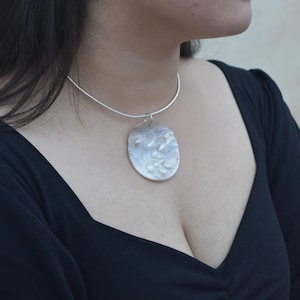 Peut inclure: Un collier en argent avec un grand pendentif rond texturé. Le pendentif a une finition blanche nacrée.