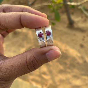 May include: Two silver rings with a heart shape design. Each ring has a red gemstone set in the center of the heart.
