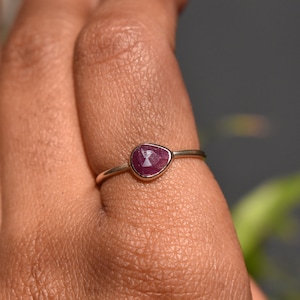 Può includere: Un anello d'argento con una pietra preziosa sfaccettata di colore rosso scuro incastonata in una fascia semplice e delicata.