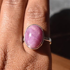 Puede incluir: Anillo de plata con una gran gema rosa ovalada. La gema tiene una superficie lisa y pulida, engastada en una sencilla banda de plata. El anillo se muestra en un dedo, destacando su tamaño y color. Joyería.