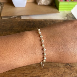 Op de afbeelding: Een delicate zilveren kettingarmband met kleine, witte, iriserende kralen. De armband zit om een pols.