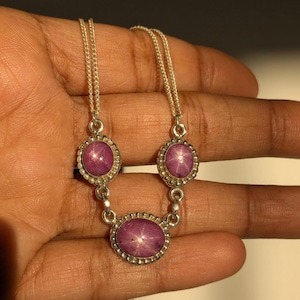 May include: A silver necklace featuring three oval-shaped, star-like ruby gemstones. The rubies are set in a silver bezel with a beaded border, connected by delicate silver chains. The necklace is displayed on a hand.