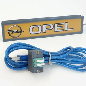 Puede incluir: Un letrero gris y amarillo con la palabra "OPEL" en letras blancas y el logotipo de Opel. El letrero est&aacute; conectado a un cable USB azul con un conector gris.