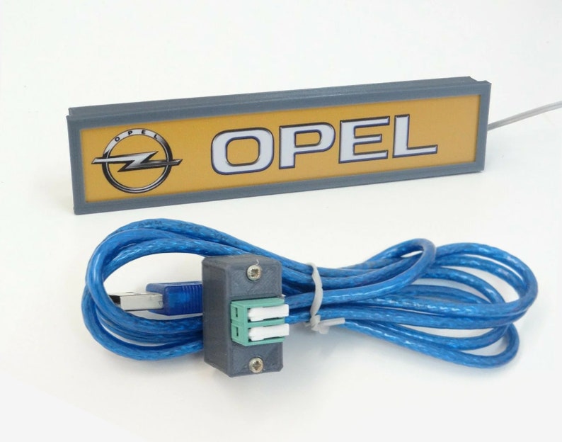 Puede incluir: Un letrero gris y amarillo con la palabra "OPEL" en letras blancas y el logotipo de Opel. El letrero est&aacute; conectado a un cable USB azul con un conector gris.