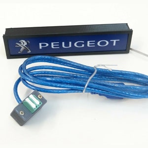 Puede incluir: Una caja rectangular negra con fondo azul y texto blanco que dice "PEUGEOT". Un logotipo de le&oacute;n plateado se encuentra en el lado izquierdo de la caja. Un cable azul con un conector gris est&aacute; conectado a la caja.