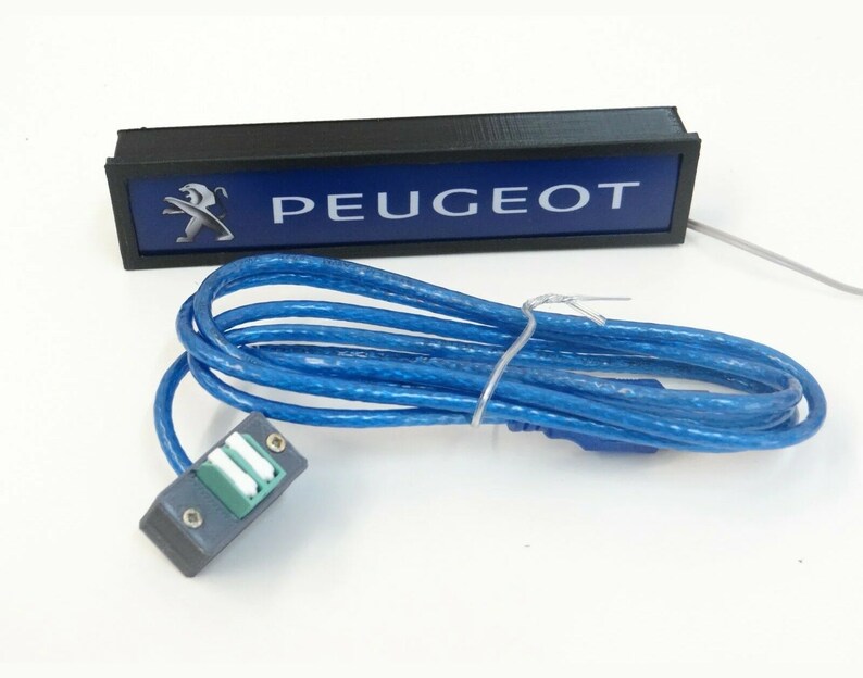 Puede incluir: Una caja rectangular negra con fondo azul y texto blanco que dice "PEUGEOT". Un logotipo de le&oacute;n plateado se encuentra en el lado izquierdo de la caja. Un cable azul con un conector gris est&aacute; conectado a la caja.