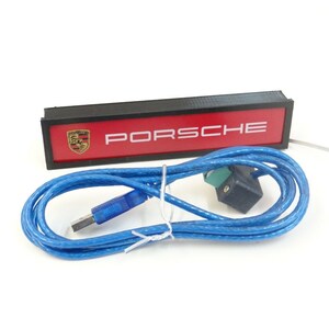 Puede incluir: Un letrero rectangular negro con fondo rojo y texto blanco que dice "PORSCHE". El letrero tiene un peque&ntilde;o logotipo en blanco y negro del escudo de Porsche en la esquina superior izquierda. El letrero est&aacute; conectado a un cable USB azul.