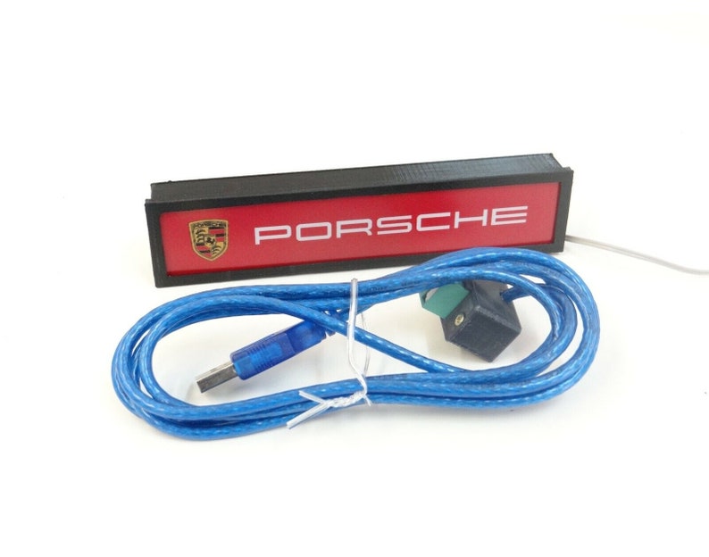 Puede incluir: Un letrero rectangular negro con fondo rojo y texto blanco que dice "PORSCHE". El letrero tiene un peque&ntilde;o logotipo en blanco y negro del escudo de Porsche en la esquina superior izquierda. El letrero est&aacute; conectado a un cable USB azul.