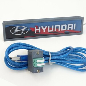 Puede incluir: Un letrero rectangular gris con la palabra "HYUNDAI" en letras blancas y un logotipo de Hyundai rojo y plateado. El letrero est&aacute; conectado a un cable USB azul con un conector gris.