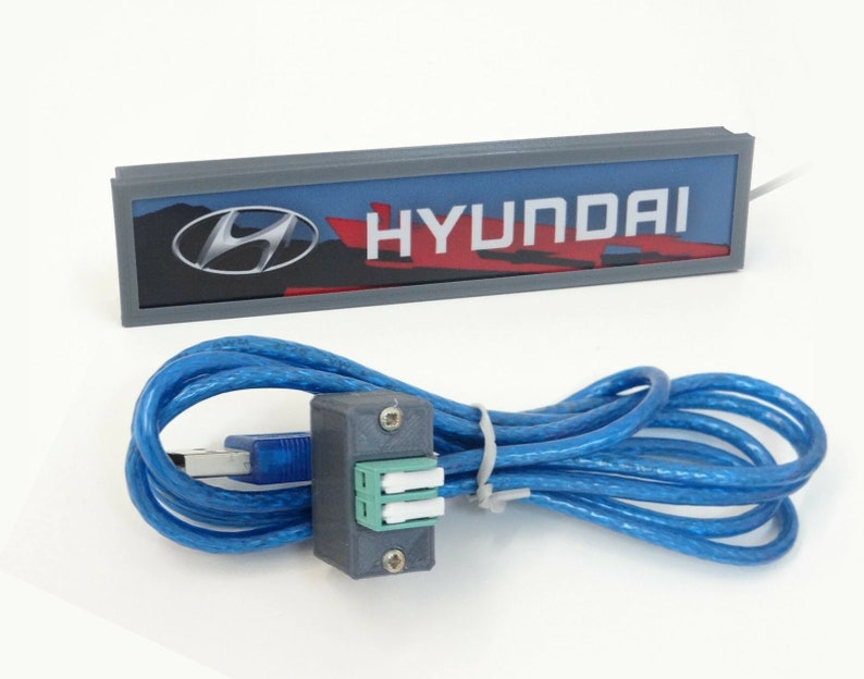 Puede incluir: Un letrero rectangular gris con la palabra "HYUNDAI" en letras blancas y un logotipo de Hyundai rojo y plateado. El letrero est&aacute; conectado a un cable USB azul con un conector gris.