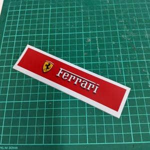Puede incluir: Una pegatina rectangular roja y blanca con el logotipo de Ferrari y la palabra "Ferrari" en letras blancas.