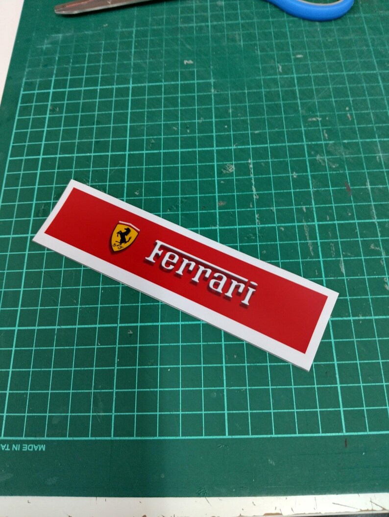 Puede incluir: Una pegatina rectangular roja y blanca con el logotipo de Ferrari y la palabra "Ferrari" en letras blancas.