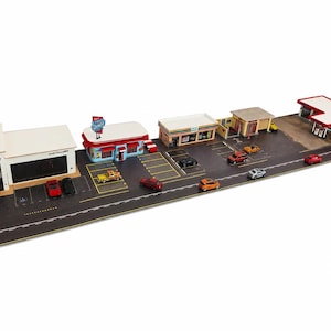 May include: A miniature diorama set featuring a car gallery, a diner with a sign, a convenience store, and a gas station. The layout includes a paved road, parking spaces, and several small toy cars in various colors arranged across the scene.