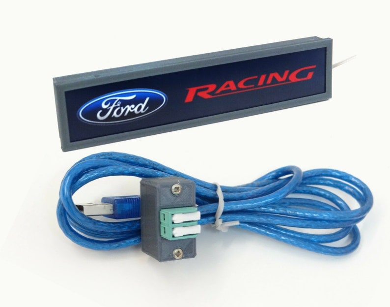Puede incluir: Un letrero rectangular gris con un logotipo Ford azul y la palabra "RACING" en letras rojas. El letrero est&aacute; conectado a un cable USB azul con un conector gris.