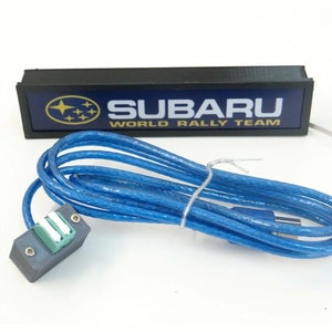 Puede incluir: Un letrero rectangular negro con fondo azul y el texto "SUBARU WORLD RALLY TEAM" en letras amarillas. El letrero tiene un logotipo Subaru blanco con estrellas doradas. El letrero est&aacute; conectado a un cable USB azul.