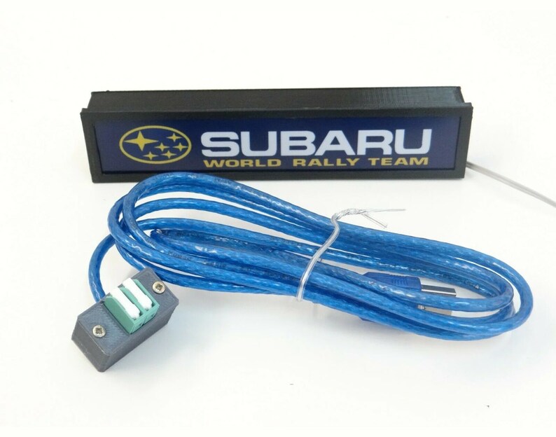 Puede incluir: Un letrero rectangular negro con fondo azul y el texto "SUBARU WORLD RALLY TEAM" en letras amarillas. El letrero tiene un logotipo Subaru blanco con estrellas doradas. El letrero est&aacute; conectado a un cable USB azul.