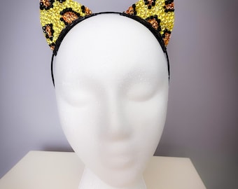 May include: A black headband with yellow and orange leopard print cat ears. The ears are made of rhinestones.