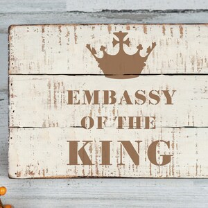 Puede incluir: Un letrero de madera blanco con letras marrones desgastadas que dice "Embassy of the King" con una corona marrón sobre el texto.