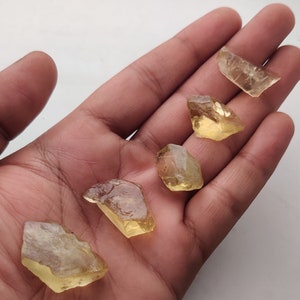May include: Five yellow-toned, faceted, rough-cut gemstones. The stones are translucent and have a slight golden hue. They are arranged on a hand palm.