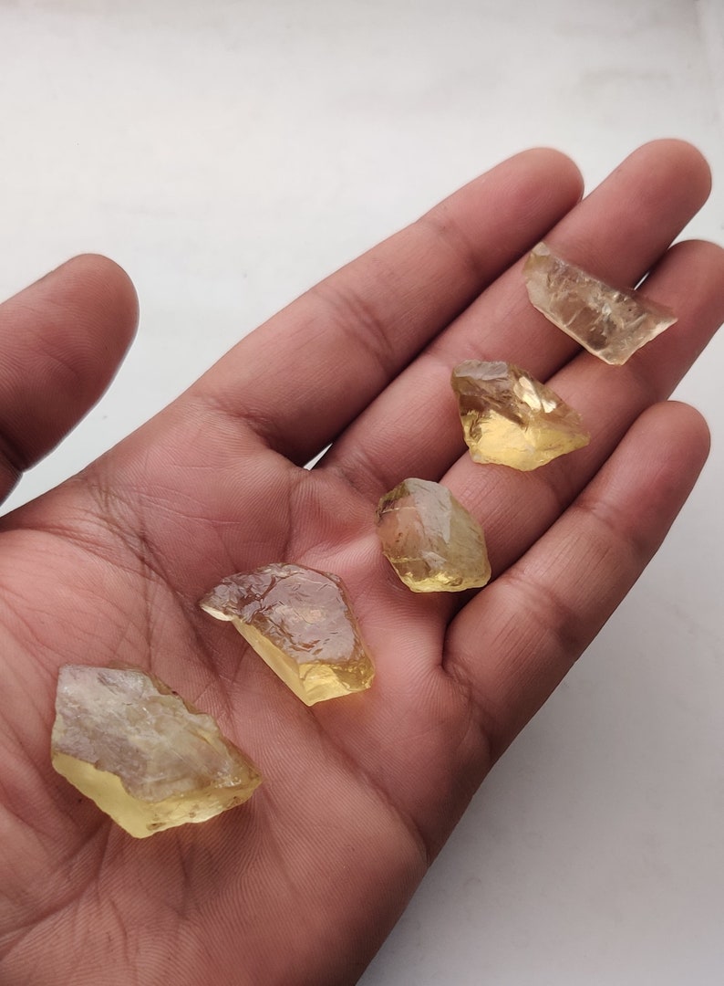 May include: Five yellow-toned, faceted, rough-cut gemstones. The stones are translucent and have a slight golden hue. They are arranged on a hand palm.