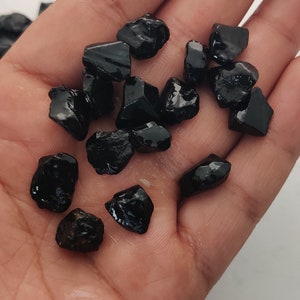 May include: A collection of black gemstone chips, possibly obsidian or black tourmaline, in various sizes and shapes. The chips are scattered on a person's hand.