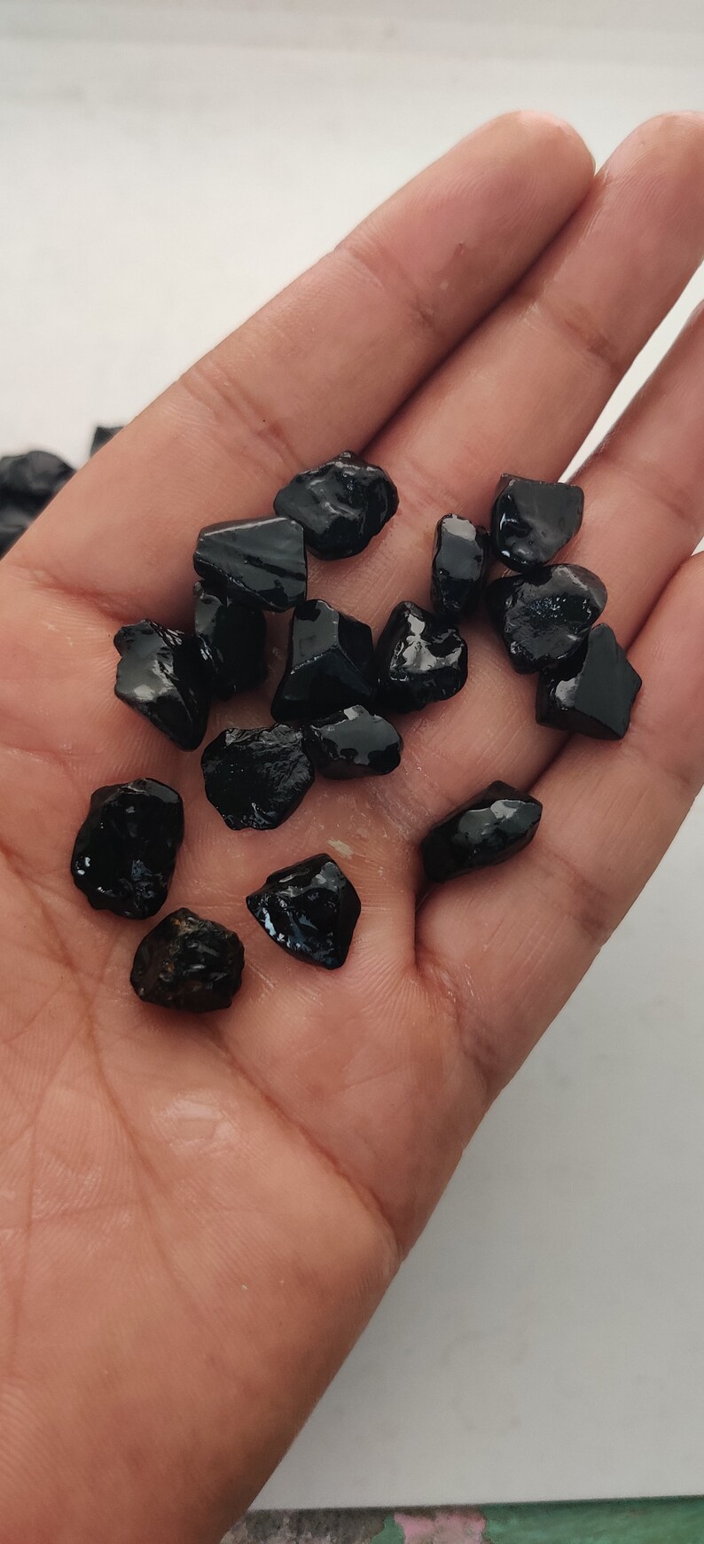 May include: A collection of black gemstone chips, possibly obsidian or black tourmaline, in various sizes and shapes. The chips are scattered on a person's hand.