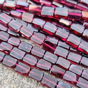 Puede incluir: Una hebra de cuentas de vidrio rectangulares de color rojo oscuro. Las cuentas son translúcidas y tienen un ligero brillo.