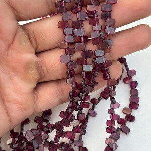 May include: Strands of dark red garnet beads. The beads are rectangular and strung together, creating a necklace or bracelet. The beads have a polished finish and vary in color from deep red to a lighter, translucent shade.