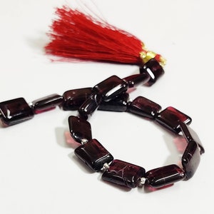 Pode incluir: Um fio de contas de pedras preciosas vermelho-escuras com um tassel vermelho. As contas são retangulares e têm um acabamento brilhante.