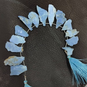 Op de afbeelding: Een streng lichtblauwe edelsteen kralen met een ruwe, onregelmatige vorm. De kralen zijn geregen op een zilveren draad en hebben aan elk uiteinde blauwe kwastjes.