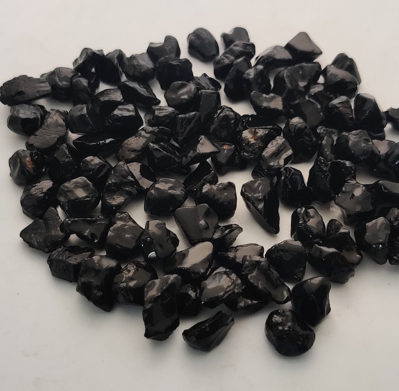 May include: A collection of black obsidian chips, a type of volcanic glass, with a smooth, polished finish. The chips are various sizes and shapes, and have a dark, opaque appearance.