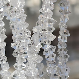 Pode incluir: Um fio de contas transparentes, facetadas, em forma de diamante. As contas são enfiadas em um fio transparente e estão dispostas em linha vertical. As contas são todas do mesmo tamanho e forma, e são muito brilhantes.