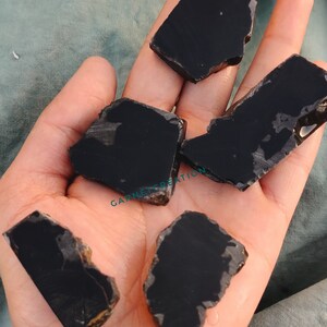 K&ouml;nnte beinhalten: F&uuml;nf St&uuml;cke schwarzer Obsidian, ein dunkles, glasartiges Vulkangestein, werden auf einer Hand gezeigt. Die St&uuml;cke sind unregelm&auml;&szlig;ig geformt und haben eine glatte, polierte Oberfl&auml;che.