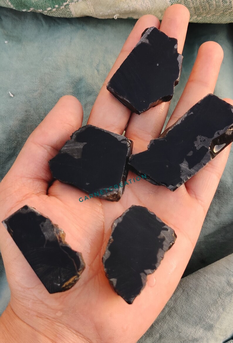 K&ouml;nnte beinhalten: F&uuml;nf St&uuml;cke schwarzer Obsidian, ein dunkles, glasartiges Vulkangestein, werden auf einer Hand gezeigt. Die St&uuml;cke sind unregelm&auml;&szlig;ig geformt und haben eine glatte, polierte Oberfl&auml;che.