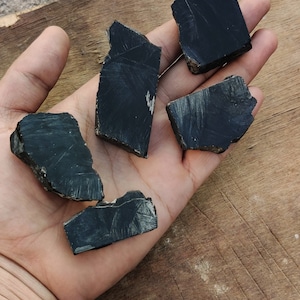K&ouml;nnte beinhalten: F&uuml;nf St&uuml;cke schwarzer Obsidian, ein dunkles, glasartiges Vulkangestein mit einer rauen, strukturierten Oberfl&auml;che. Der Obsidian ist auf einer Holzoberfl&auml;che angeordnet.