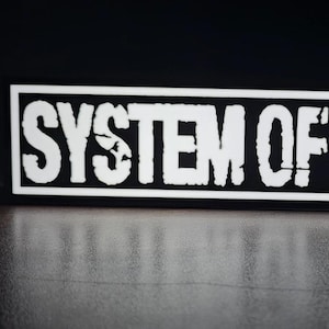 May include: A rectangular black and white sticker with the text "SYSTEM OF A DOWN" in a bold, distressed font. The text is white, and the sticker has a black border. The sticker is on a reflective surface.