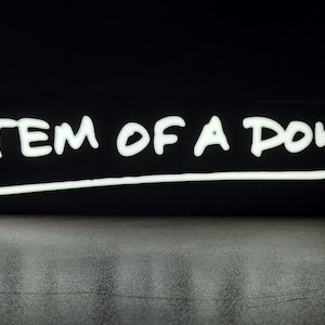 May include: Black rectangular object with the white text "SYSTEM OF A DOWN" in a handwritten style, underlined with a white line. The background is black, and the object is on a reflective surface.