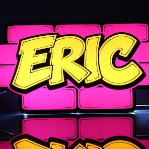 May include: A neon sign featuring the name "ERIC" in a vibrant yellow, outlined in black, against a backdrop of pink brick-like shapes. The sign is illuminated, creating a bright and eye-catching display.