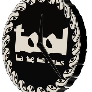 Op de afbeelding: Een zwart-witte klok met het woord "Tool" in een gestileerd lettertype en het woord "Lateralus". De klok heeft een decoratieve rand en zwarte wijzers. De klok heeft een ronde vorm.