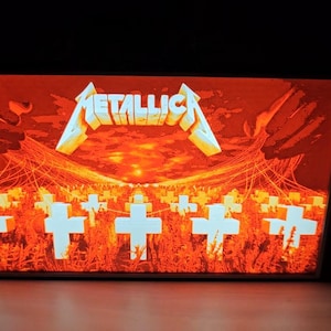 Può includere: Immagine incorniciata con il logo Metallica in bianco e oro su uno sfondo arancione infuocato. L'opera raffigura un campo di croci bianche sotto un tramonto, con il nome della band in evidenza.