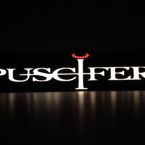 May include: A black and white sign with the word "Puscifer" in white letters and a red crescent moon above a black vertical line.