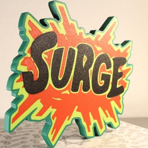 May include: A three-dimensional sign with the word "SURGE" in black, set against a red and yellow burst design. The sign has a green outline and is likely made of wood or a similar material. The sign is displayed on a patterned surface.
