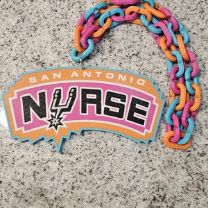 Pode incluir: Uma placa decorativa com o texto SAN ANTONIO NURSE e um logótipo de espora inspirado no basquetebol a substituir a letra U. A placa está ligada a uma corrente de plástico multicolorida com elos alternados em laranja, rosa e azul claro.