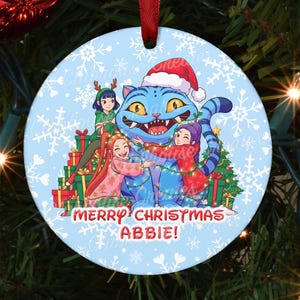 May include: A round Christmas ornament with a light blue background and a cartoon illustration. The illustration features a blue cat wearing a Santa hat, surrounded by children and Christmas presents. The text "MERRY CHRISTMAS ABBIE!" is written on the ornament.