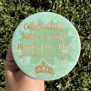 May include: A green round sign with a gold glitter finish and gold lettering that reads "Celebrating Justine & Cody's Happily Ever After! January 11th, 2022". A gold glitter crown is in the bottom center of the sign.