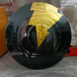 Op de afbeelding: Een rond zwart schild met een gouden bliksemontwerp. Het schild is gemaakt van hard materiaal en heeft een glanzende afwerking.