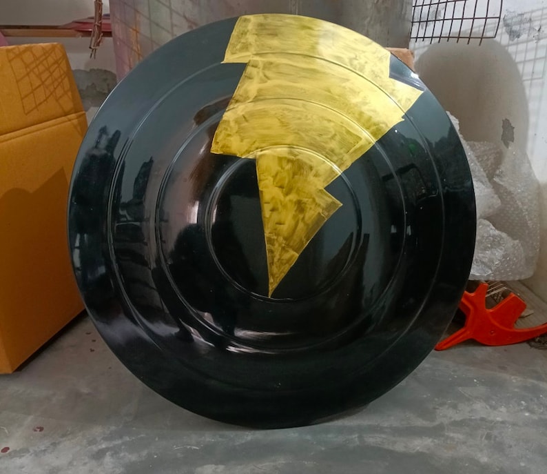 Op de afbeelding: Een rond zwart schild met een gouden bliksemontwerp. Het schild is gemaakt van hard materiaal en heeft een glanzende afwerking.