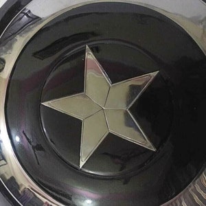 Puede incluir: Un escudo circular negro y plateado con una estrella de cinco puntas en el centro. La estrella es de plata y tiene una superficie reflectante.